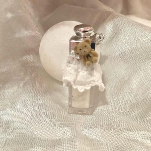 May include: A small, clear rectangular perfume bottle with a silver cap and a tiny teddy bear attached. The bottle is adorned with a white lace trim. The background is a white, textured fabric.