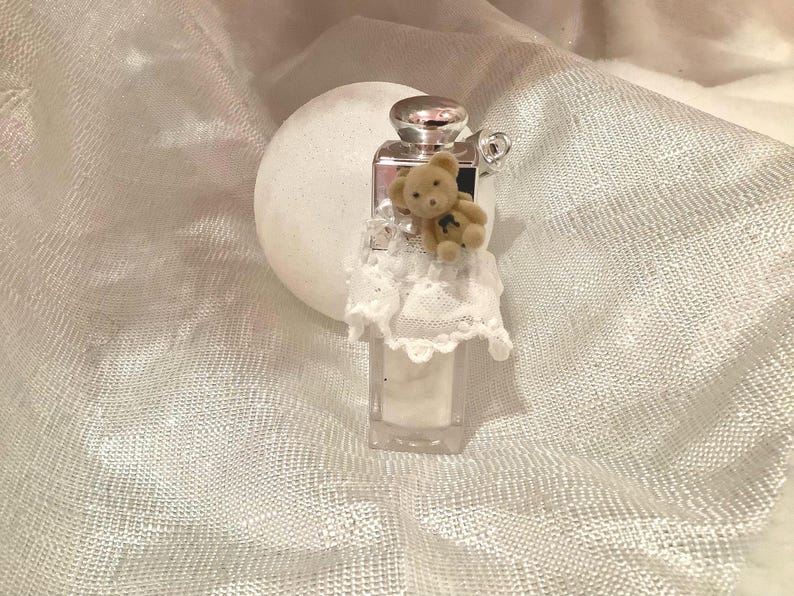 May include: A small, clear rectangular perfume bottle with a silver cap and a tiny teddy bear attached. The bottle is adorned with a white lace trim. The background is a white, textured fabric.