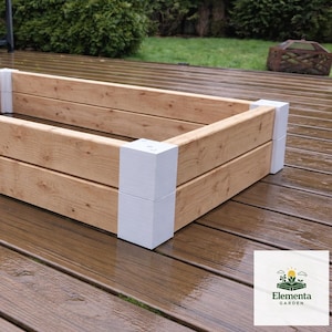 May include: A rectangular wooden raised garden bed with light brown planks and white corner posts. The garden bed is empty and sits on a wooden deck. The Elementa Garden logo is visible in the bottom right corner.