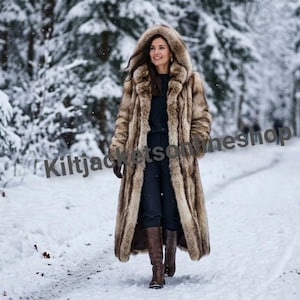 May include: A long, hooded faux fur coat in shades of brown and tan, worn over a dark top and trousers with brown leather boots. The text Kiltjacketsonlineshop is overlaid across the center of the image, which is set in a snowy forest.