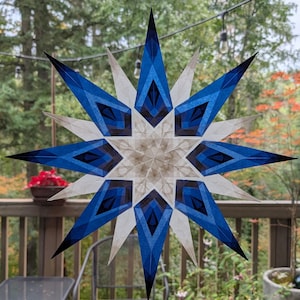 Op de afbeelding: Een stervormig ornament met meerdere punten en een geometrisch ontwerp met afwisselende lagen van diepblauw en doorschijnend wit papier. De ster vertoont een complex, symmetrisch patroon met scherpe, langwerpige punten vanuit het midden.