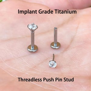 May include: Three silver-colored, threadless push pin studs. Each stud has a small, clear, round gemstone. The text "Implant Grade Titanium" and "Threadless Push Pin Stud" are displayed in black.