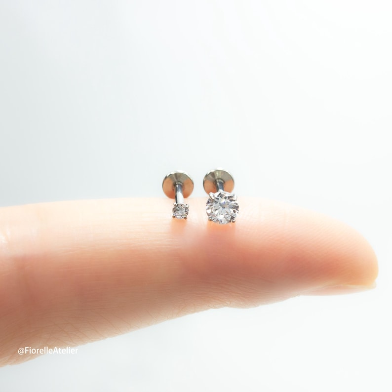May include: Two silver stud earrings with clear, round-cut gemstones. One earring features a smaller, square-cut stone, while the other has a larger, more prominent round stone. The earrings are displayed on a light-colored surface.