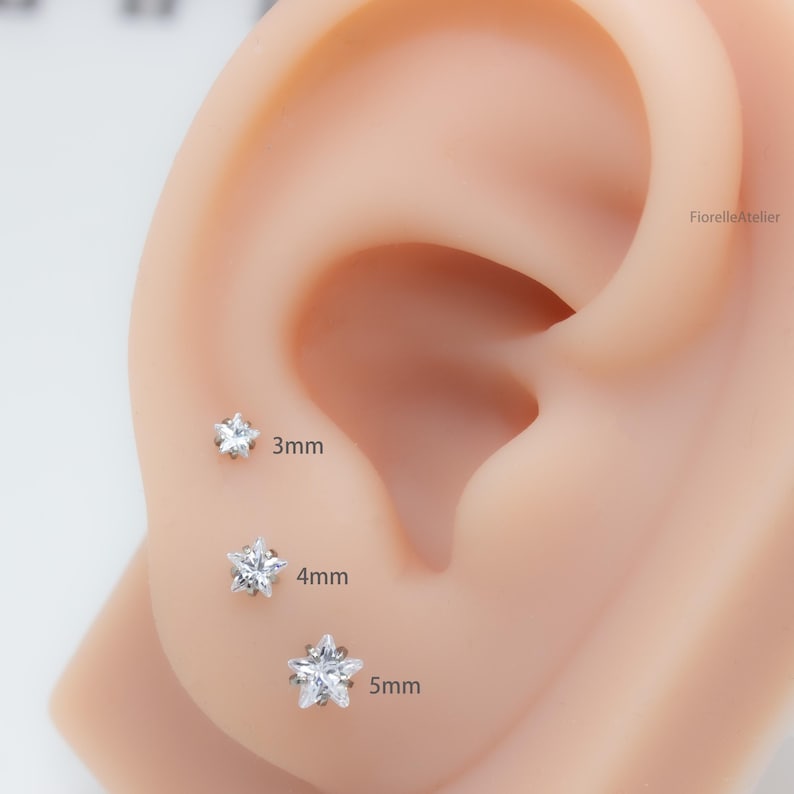 Może przedstawiać: Trzy kolczyki w kształcie gwiazdki z przezroczystymi kamieniami szlachetnymi są prezentowane na uchu. Kolczyki mają rozmiar 3 mm, 4 mm i 5 mm. Kolczyki są umieszczone na jasnym tle.