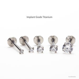 May include: Five silver-colored, implant-grade titanium labret studs with clear, round cubic zirconia stones. The text "Implant Grade Titanium" is above the studs. The studs vary in size, with the largest stone on the right.