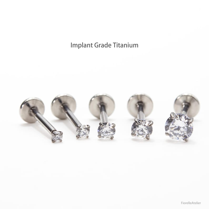 May include: Five silver-colored, implant-grade titanium labret studs with clear, round cubic zirconia stones. The text "Implant Grade Titanium" is above the studs. The studs vary in size, with the largest stone on the right.