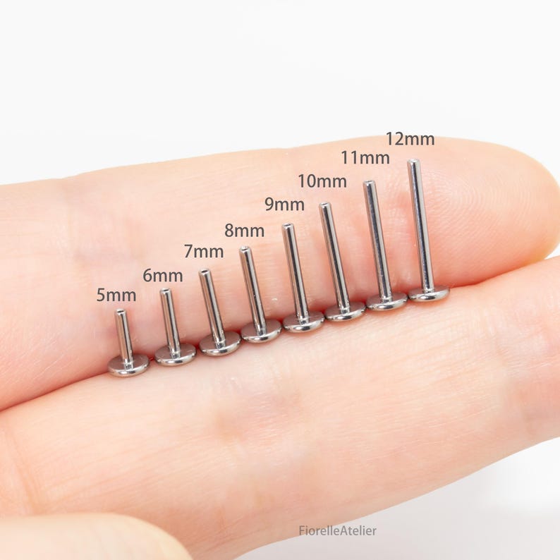 May include: A collection of silver-colored lip piercing studs, ranging in size from 5mm to 12mm. Each stud features a flat disc base and a straight post. The sizes are labeled in millimeters, with the largest stud measuring approximately 0.47 inches.