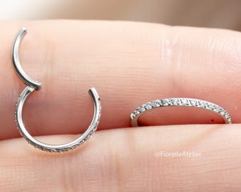 Peut inclure: Deux boucles d'oreilles créoles argentées articulées, serties de petits pierres précieuses transparentes le long de la courbe extérieure. Le texte "Implant-Grade Titanium" est visible en haut. Le nom de la marque "@FiorelleAtelier" est en bas.