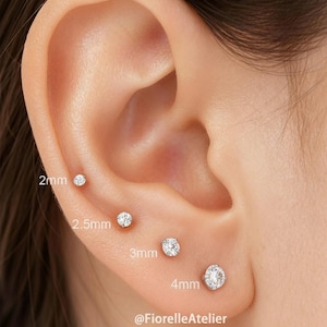 May include: Close-up of an ear with four small, round, clear stud earrings of varying sizes: 2mm, 2.5mm, 3mm, and 4mm. The earrings are set in the earlobe and helix. The image includes the text "@FiorelleAtelier".