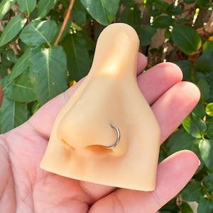 May include: A tan-coloured nose model featuring a silver nose ring. The model is held in a hand, with green foliage in the background. The nose ring is a simple, circular design.