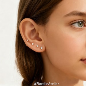 May include: Close-up of an ear with four small, round, diamond stud earrings. The earrings are set in gold and increase in size from top to bottom. The skin tone is fair, and the background is blurred.