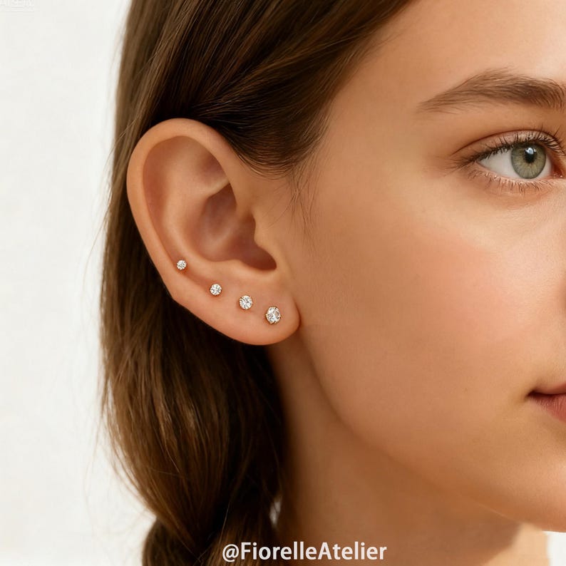 May include: Close-up of an ear with four small, round, diamond stud earrings. The earrings are set in gold and increase in size from top to bottom. The skin tone is fair, and the background is blurred.