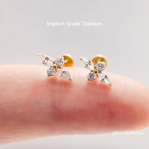May include: A pair of gold-colored stud earrings, each featuring a four-petal flower design. Each petal is a marquise-cut clear stone, with a round clear stone in the center. The text "Implant-Grade Titanium" is visible.