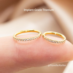 May include: Two gold-colored, circular rings with clear gemstones set along the outer edge. The rings are held on a finger. The text "Implant-Grade Titanium" is above the rings. The rings are likely body jewelry.
