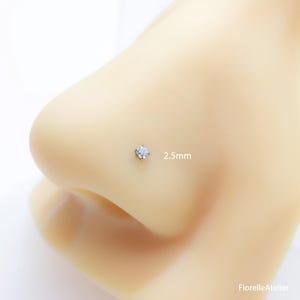 May include: A small, silver nose stud with a clear, square-cut gemstone. The stud is 2.5mm in size and is set on a light-colored nose. The jewelry is from FiorelleAtelier.