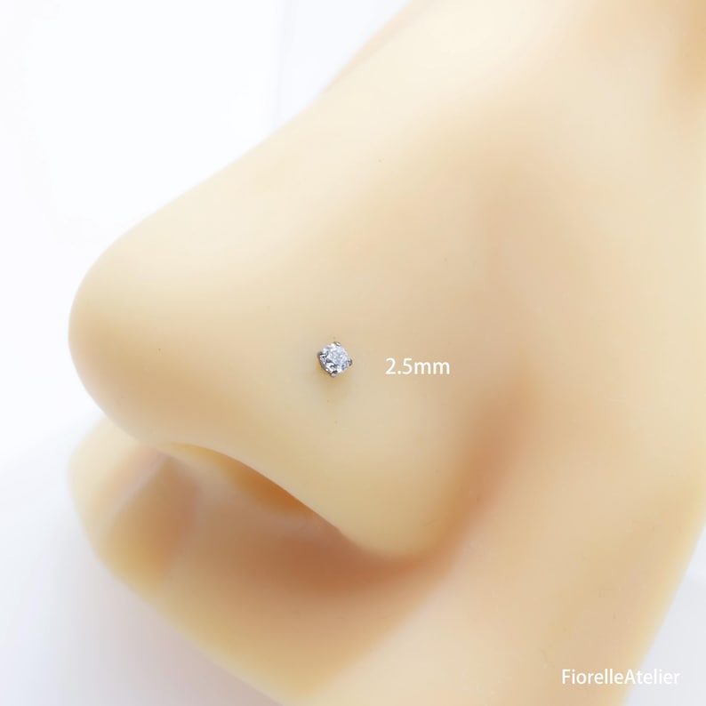 May include: A small, silver nose stud with a clear, square-cut gemstone. The stud is 2.5mm in size and is set on a light-colored nose. The jewelry is from FiorelleAtelier.
