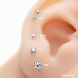 May include: A close-up of an ear with five clear, round-cut stud earrings of varying sizes: 1.5mm, 2mm, 2.5mm, 3mm, and 4mm. The earrings are set in silver or white metal. The text "FiorelleAtelier" is in the bottom right corner.