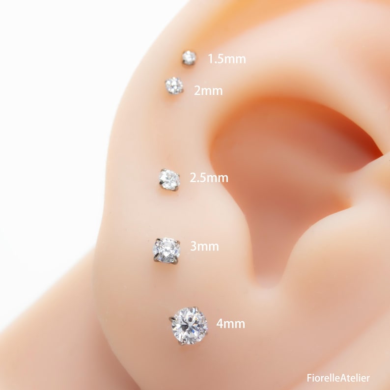 May include: A close-up of an ear with five clear, round-cut stud earrings of varying sizes: 1.5mm, 2mm, 2.5mm, 3mm, and 4mm. The earrings are set in silver or white metal. The text "FiorelleAtelier" is in the bottom right corner.