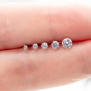 May include: A selection of six clear, round-cut diamond stud earrings of varying sizes, displayed in a row. The earrings are set in a silver metal. The image is set against a soft, neutral background.
