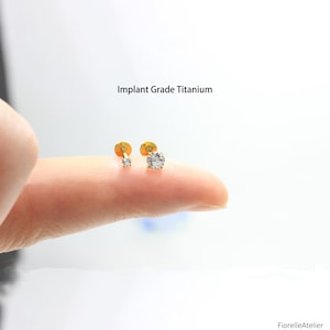 May include: Two gold-tone stud earrings featuring clear, round-cut stones in a prong setting. The text Implant Grade Titanium is visible above the jewelry. The earrings are positioned on a finger to show scale, with a flat back post design.