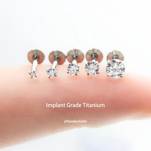 May include: Five silver-colored, implant-grade titanium stud earrings with clear, round-cut gemstones of varying sizes. The text "Implant Grade Titanium" is displayed below the earrings.