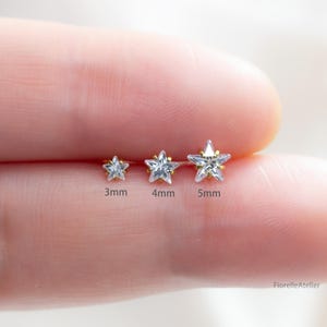 May include: Three gold star-shaped earrings with clear cubic zirconia stones. The earrings are 3mm, 4mm, and 5mm in size. The image shows the earrings on a person's finger.