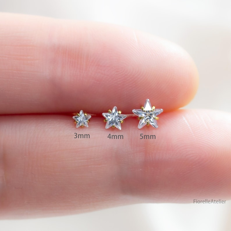 Może przedstawiać: Trzy złote kolczyki w kształcie gwiazdki z przezroczystymi kamieniami z cyrkonii sześciennej. Kolczyki mają 3 mm, 4 mm i 5 mm. Zdjęcie przedstawia kolczyki na palcu osoby.