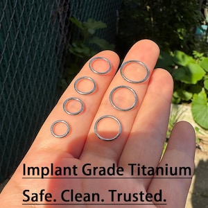 May include: Several silver-coloured, circular body jewellery rings of different sizes. The text "Implant Grade Titanium. Safe. Clean. Trusted." is displayed below the rings.