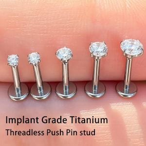 May include: Five silver threadless push pin studs with clear gemstones. The studs are displayed in a row, with the gemstones increasing in size. The text "Implant Grade Titanium Threadless Push Pin stud" is visible.