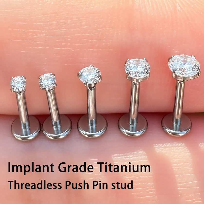 May include: Five silver-colored, threadless push pin studs with clear, sparkling gemstones. The studs are arranged in a row, showcasing varying sizes of the gemstones. The text "Implant Grade Titanium Threadless Push Pin stud" is visible.