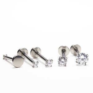 May include: A collection of silver-colored lip or ear piercing studs. The studs have a flat disc backing and a post with either a small, clear gemstone or a flat, round disc on the front. The background is white.