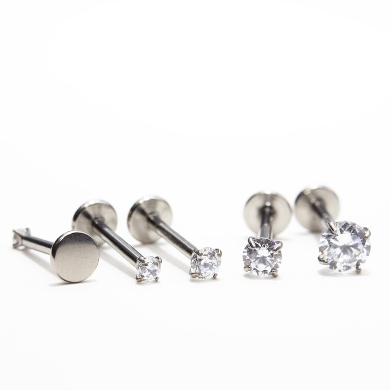 May include: A collection of silver-colored lip or ear piercing studs. The studs have a flat disc backing and a post with either a small, clear gemstone or a flat, round disc on the front. The background is white.
