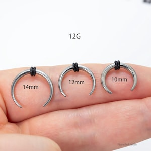 Peut inclure: Trois bijoux de corps argent&eacute;s en forme de croissant avec des bandes de caoutchouc noires. Les tailles sont de 14 mm, 12 mm et 10 mm. Le texte "12G" est au-dessus des bijoux.
