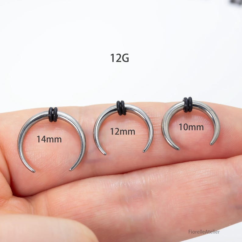 Peut inclure: Trois bijoux de corps argent&eacute;s en forme de croissant avec des bandes de caoutchouc noires. Les tailles sont de 14 mm, 12 mm et 10 mm. Le texte "12G" est au-dessus des bijoux.