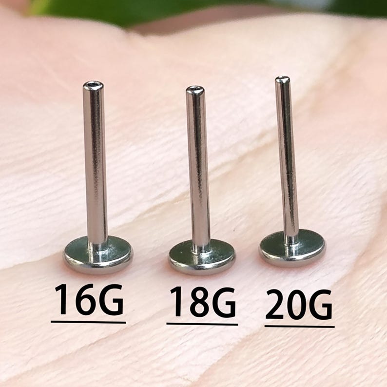 May include: Three silver lip piercing studs with flat disc backs. The gauges are labeled 16G, 18G, and 20G. The posts are cylindrical and have a small opening at the top. The background is a soft, blurred pink.