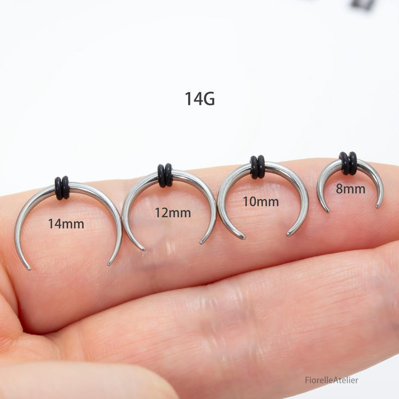 Peut inclure: Quatre bijoux de corps argent&eacute;s en forme de fer &agrave; cheval avec des anneaux en caoutchouc noirs. Les tailles sont de 14 mm, 12 mm, 10 mm et 8 mm. Le texte "14G" est au-dessus des bijoux.