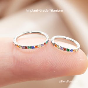 May include: Two silver-colored, circular nose rings with a row of colorful gemstones. The rings are made of implant-grade titanium. The text "Implant-Grade Titanium" is visible in the image.