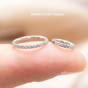 May include: Two small, silver-colored rings with clear gemstones are displayed. The rings are set with multiple small, clear stones. The text "Implant-Grade Titanium" is visible above the rings. The rings are on a light-colored background.