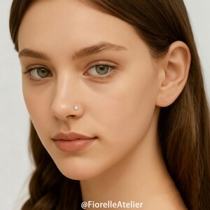 May include: A close-up of a person's face with a small, sparkling, clear gemstone nose stud. The stud is set in a silver or white metal. The skin tone is fair, and the eyes are green. The image is well-lit, highlighting the jewelry.
