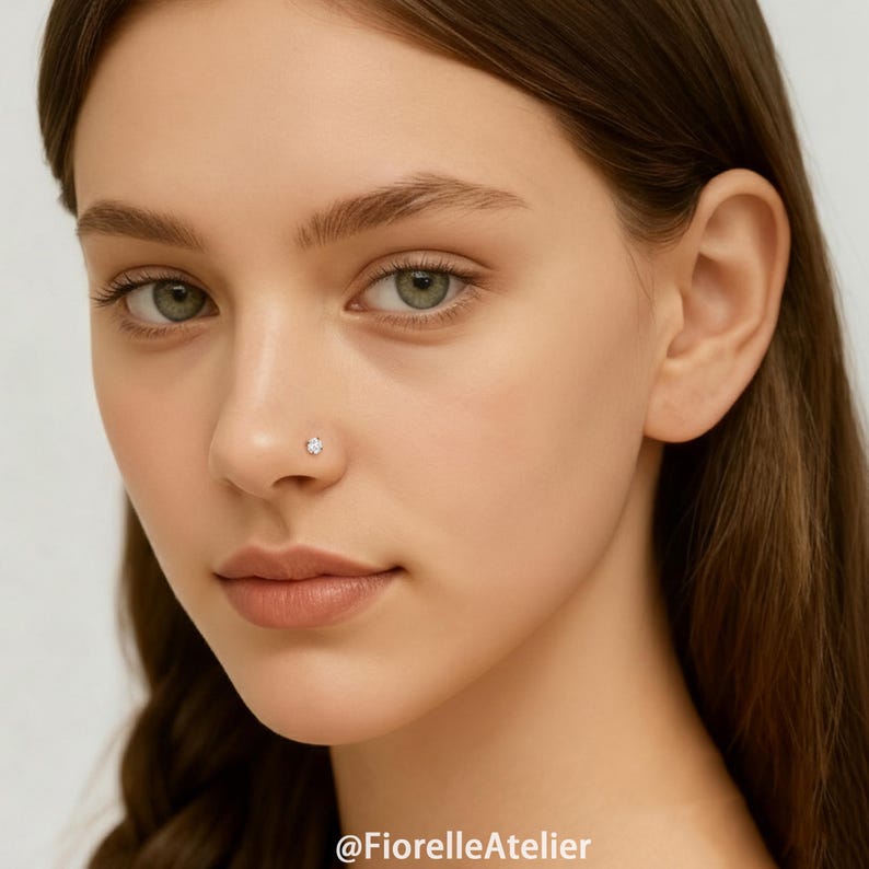 May include: A close-up of a person's face with a small, sparkling, clear gemstone nose stud. The stud is set in a silver or white metal. The skin tone is fair, and the eyes are green. The image is well-lit, highlighting the jewelry.