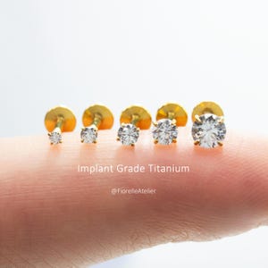 May include: Five gold-tone stud earrings with clear round-cut stones in varying sizes, arranged in a row. The text Implant Grade Titanium and @FiorelleAtelier is visible. Each earring features a prong setting and a flat back post design.