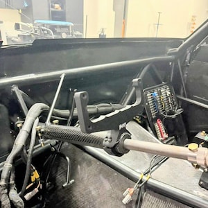 May include: Interior view of a race car cockpit. The image shows a black steering wheel with a carbon fiber grip, a black metal handle, and various wiring components. The car's interior is primarily black.