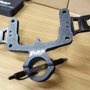 May include: A black and gray mechanical device with the text "WMRP" in white. A black pen is held in a circular component. The device has multiple rectangular and circular openings. The device is on a wooden surface.