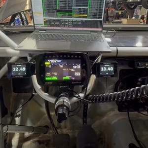 May include: A car's dashboard with multiple digital displays showing data. A laptop sits above the dashboard, displaying data. The displays show numbers like 12.49, 13.40, and 1187. The dashboard is mounted on a white metal frame.