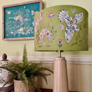 Op de afbeelding: Een tafellamp met een cilindervormige voet van licht hout en een olijfgroene trommelvormige lampenkap met een print van witte vogels, roze nachtvlinders en diverse kleurrijke bloemelementen in een botanisch patroon.