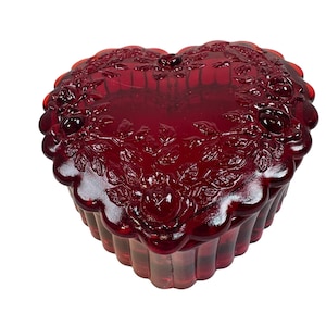 Puede incluir: Una caja de baratijas de vidrio rojo rubí en forma de corazón. La tapa y los lados presentan un diseño floral en relieve. La caja tiene un borde festoneado y está hecha de vidrio transparente. La caja probablemente se usa para guardar joyas o pequeños recuerdos.