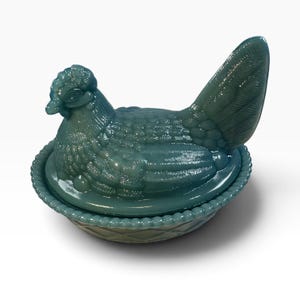 Puede incluir: Un plato de cristal color verde azulado con forma de gallina sobre un nido. La tapa tiene forma de gallina sentada con texturas de plumas detalladas, mientras que la base presenta un borde de cuentas y un patrón de cesta. El cristal es semitranslúcido.
