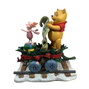May include: A decorative figurine featuring Winnie the Pooh and Piglet on a snowy train track. Winnie the Pooh, in a red shirt, holds a scroll, while Piglet stands on presents. The base is decorated with greenery, a red bow, and blue train wheels.