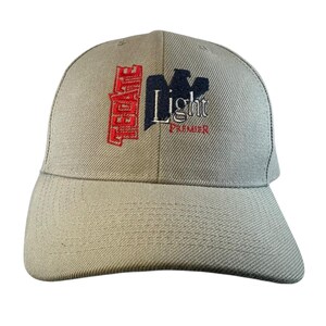May include: A tan baseball cap with the embroidered text "Tecate Light Premier" in red, navy blue, and white. The cap has a curved brim and a structured crown, suitable for outdoor wear.