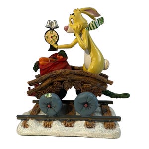 Puede incluir: Una figura de conejo amarillo con una bufanda verde y blanca, sosteniendo un pequeño reloj. El conejo está en un carro de madera con un saco rojo de zanahorias. El carro tiene ruedas azules y se asienta sobre una base nevada con vías de tren.